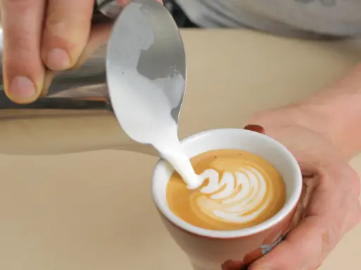 Barista gießt cremigen Milchschaum in eine Tasse Kaffee. Der Cappuccino Milchschaum bildet ein schönes Muster.