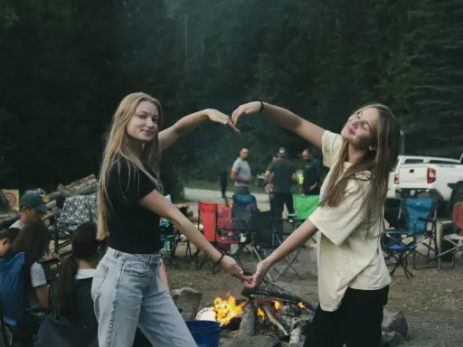 Dwie dziewczyny tworzą serce z dłoni na tle ogniska i lasu. To idealny okres na wakacjach, pełen radości i wspólnych chwil.
