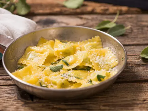 Ravioli ricotta e spinaci dorati, conditi con parmigiano e foglie di salvia fresca, serviti in una ciotola metallica su un tavolo di legno rustico.
