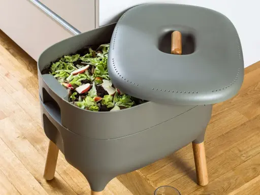 Compostador gris con restos de comida y hojas. Aprende como hacer compost para un jardín sostenible.