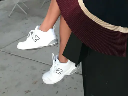 White New Balance dad shoes with a black "N" logo, paired with a pleated skirt.