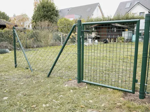 Ein grüner Maschendrahtzaun wird gespannt, mit einem Tor, das offen steht. Im Hintergrund sind Häuser und ein Schuppen zu sehen.