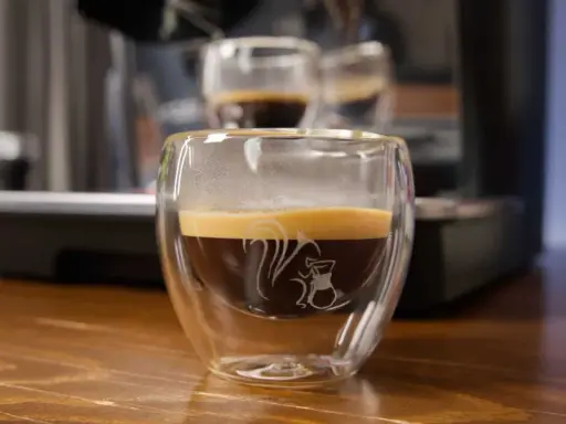Ein dampfender doppio macchiato in einem doppelwandigen Glas mit einem Eichhörnchen-Motiv.