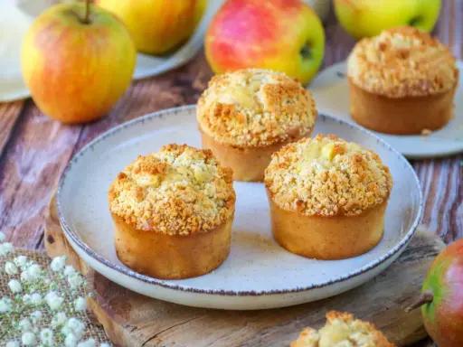 Trois muffins pomme amande dorés avec un crumble croustillant, entourés de pommes fraîches sur une table en bois.