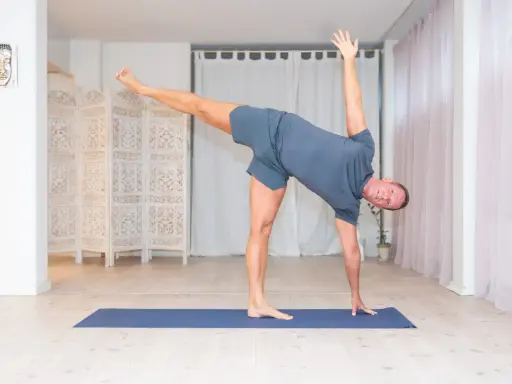 Man utför en dynamisk yoga-position på en blå matta i ett ljust rum med vita draperier och en vikbar rumsavdelare.