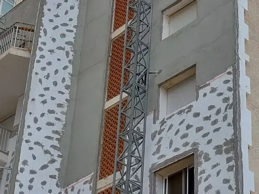 Proceso de como aislar una pared ya construida con placas de poliestireno y mortero. Se observa una grúa de obra.