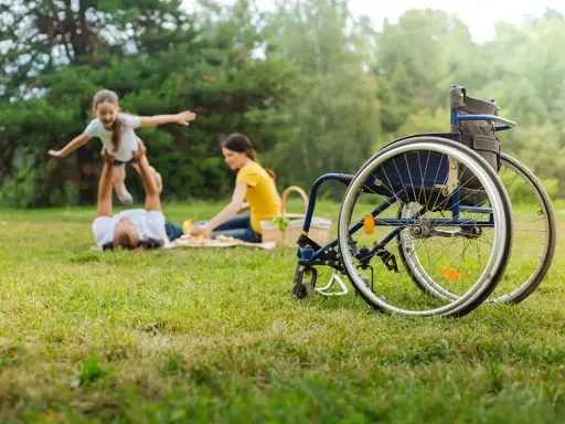 Rodzina na pikniku. Ojciec podrzuca córkę, matka przygotowuje jedzenie. Obok stoi wózek inwalidzki, symbol wsparcia i obecności opiekuna osoby niepełnosprawnej.