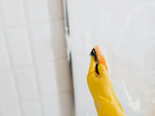 Ręka w żółtej rękawiczce czyści szklaną kabinę prysznicową gąbką. Dowiedz się, czym umyć kabinę prysznicową ze szkła, by lśniła czystością.