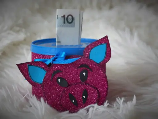 Urocza skarbonka diy w kształcie różowej świnki z brokatem, z niebieskimi uszami i kokardką. W środku widać banknot 10 zł.