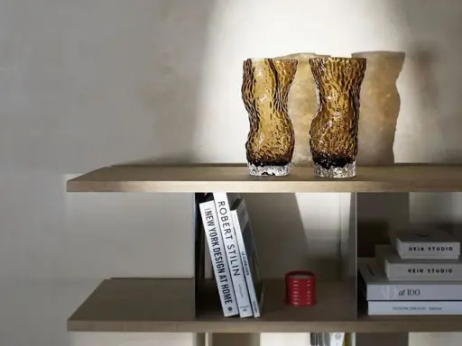 Två bärnstensfärgade glasvaser på en hylla, bredvid böcker och en röd ljushållare. Även böcker med texten HEIN STUDIO syns.