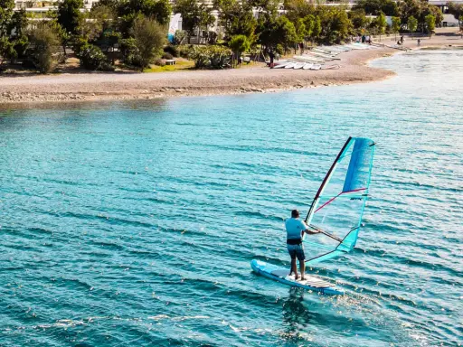 Mężczyzna na desce z żaglem pływa po turkusowej wodzie. Na brzegu widać plażę i drzewa.