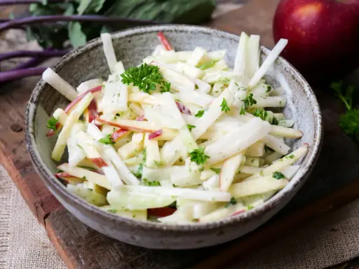 Kohlrabi Salat mit Apfelstücken und frischer Petersilie, garniert mit einem roten Apfel im Hintergrund.