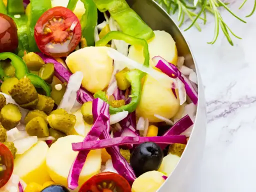 Ensalada campera vibrante con patatas, tomates, pepinillos, pimientos verdes, cebolla morada y aceitunas negras en un bol metálico.