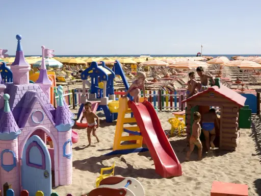 Bambini giocano in un'area giochi colorata sulla sabbia, con castello e scivoli, mentre ombrelloni gialli e lettini creano un'atmosfera da **sunday beach Rimini**.