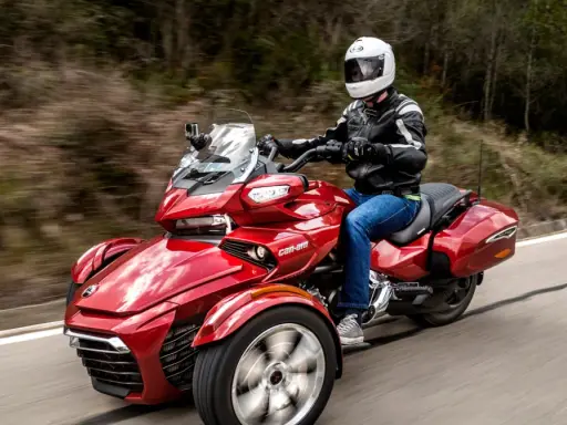 Czerwony pojazd trójkołowy z kierowcą w kasku i kurtce, jadący po krętej drodze. Idealny dla tych, którzy szukają wrażeń, ale nie mają prawa jazdy na motocykl.