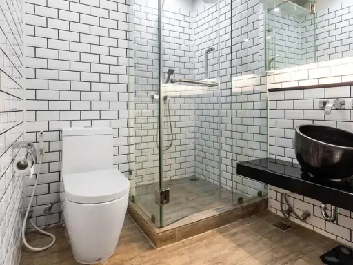 Baño moderno con azulejos blancos y negros. El suelo de madera se ve primero, luego los azulejos. Inodoro, ducha de cristal y lavabo negro.