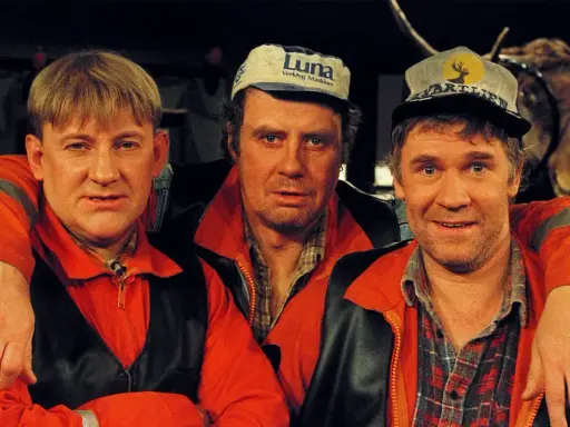 Tre män i orangea jackor och kepsar, en av dem med en uppstoppad älg i bakgrunden. Rollistan i Pistvakt.