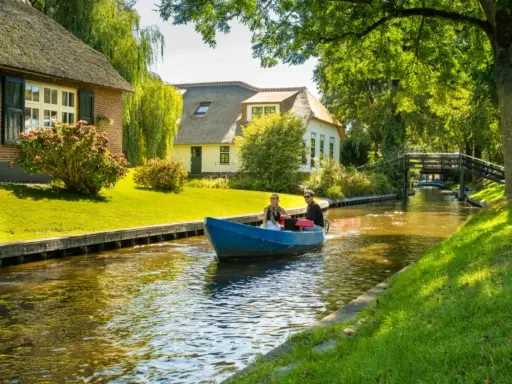 Ein Paar fährt mit einem blauen Boot durch einen Kanal im "Klein Venedig Holland". Reetgedeckte Häuser und grüne Ufer säumen den Weg.
