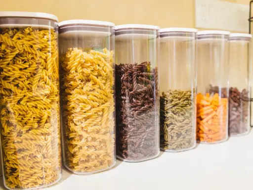 Contenitori trasparenti pieni di pasta a forma di farfalline, di vari colori: gialle, marroni, verdi e arancioni.
