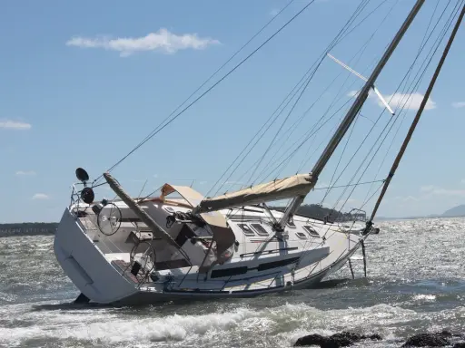 Un voilier échoué, incliné sur le côté, témoigne d'une possible collision bateau. Les vagues s'écrasent sur sa coque blanche sous un ciel bleu.