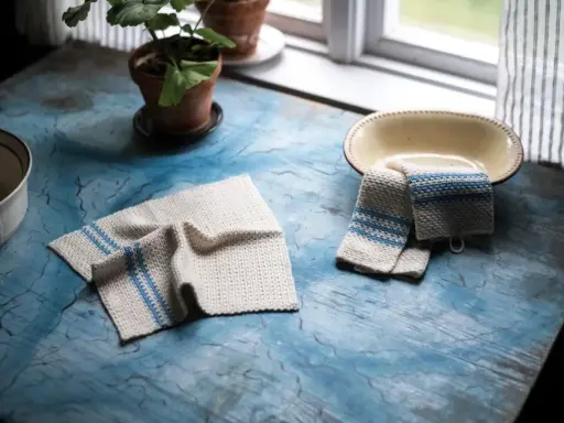 Två virka handdukar med blå ränder ligger på ett blått bord. En krukväxt och en skål syns också.