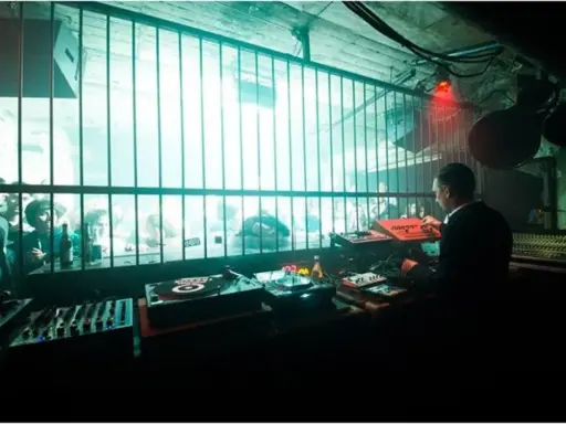 DJ set in un locale underground, luci al neon e folla che balla dietro le sbarre. Atmosfera da berlino rave.