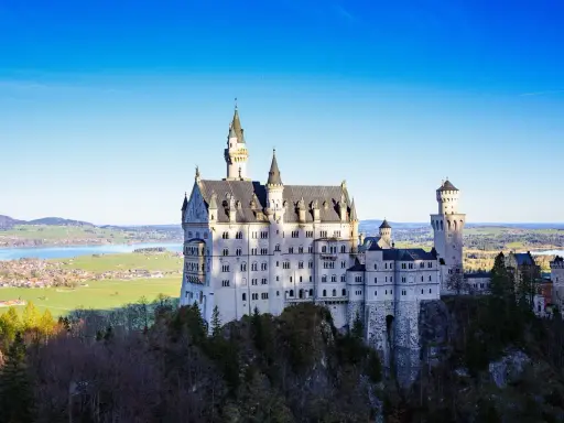 Sagolikt slott i Tyskland, Neuschwanstein, reser sig majestätiskt på en klippa med en sjö och grönskande landskap i bakgrunden.