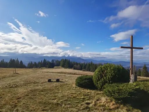 Krawcowy Wierch wita słonecznym niebem i chmurami. Drewniany krzyż góruje nad polaną, a w oddali widać pasmo górskie.
