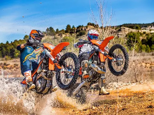 Dos pilotos de enduro compiten en un terreno embarrado, uno con motor 2t y otro 4t, salpicando agua.