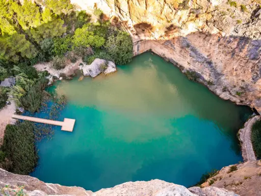 Un **charco azul** en Chiva, rodeado de rocas y vegetación. Un muelle de madera se adentra en sus aguas turquesas.