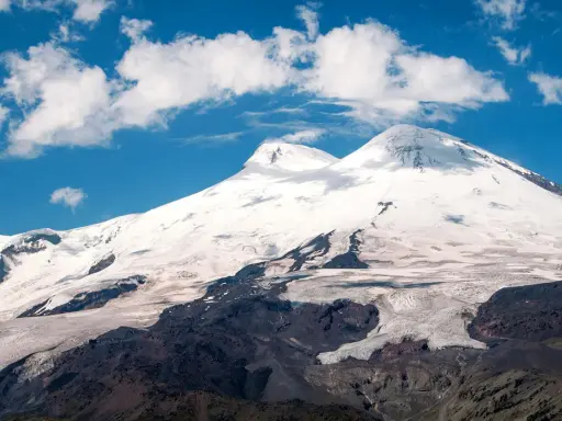 Elbrus, najwyższy szczyt Rosji, wznosi się dumnie pod błękitnym niebem z kłębiastymi chmurami. Lodowce spływają po jego zboczach.