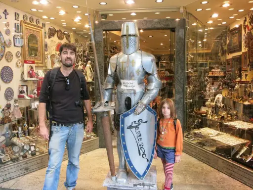 Un padre y su hija posan junto a una armadura de caballero en una tienda de souvenirs en Toledo con niños.