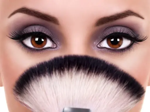 Occhi grandi e tondi esaltati da un trucco smokey viola e ciglia finte, con un pennello da trucco in primo piano.