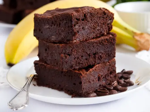 Stos puszystych brownie z bananami, ozdobiony ziarnami kawy, na talerzu z bananem w tle.