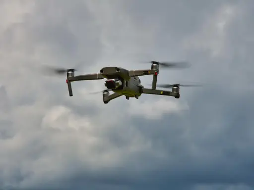 Eine graue Drohne mit roten Lichtern fliegt im bewölkten Himmel. Man kann die Propeller sehen, die sich drehen, während die Drohne in der Luft schwebt. Es ist ein tolles Gefühl, eine Drohne fliegen zu lassen.