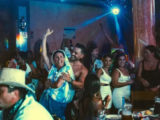 Gente celebrando y bailando en una fiesta, con luces de discoteca y un ambiente festivo. Parece una despedida de soltero con juegos y mucha alegría.