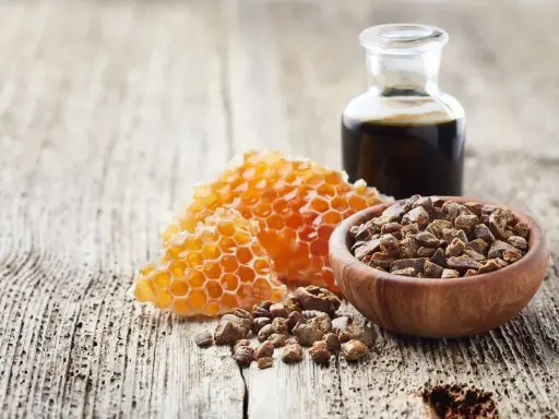 Propolis i pyłek pszczeli w drewnianej miseczce, obok plastra miodu i buteleczki z ciemnym płynem, na drewnianym tle.