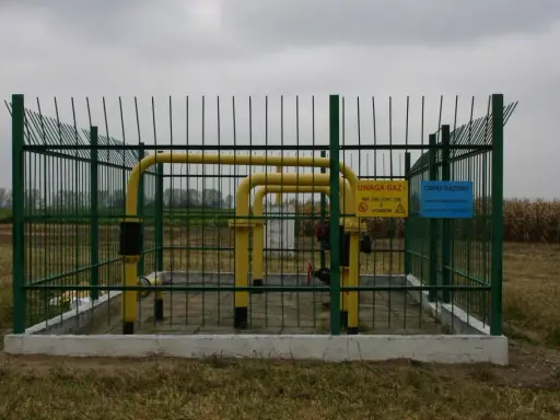 Ogrodzony obiekt gazowy z żółtymi rurami. Tablice ostrzegawcze informują o zagrożeniu. Widać pola uprawne w tle, co sugeruje odległość budynku od gazociągu średniego ciśnienia.