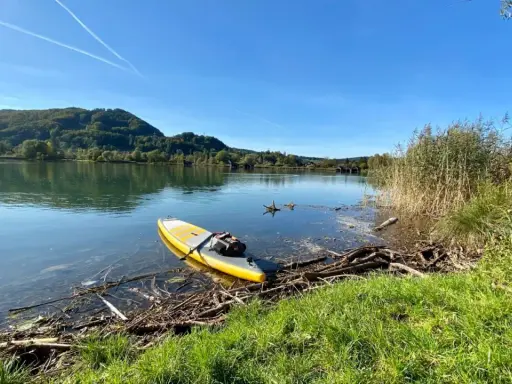 Gelbes SUP-Board liegt am Ufer bereit für SUP Touren Bayern. Ruhiger See, bewaldete Hügel und blauer Himmel.