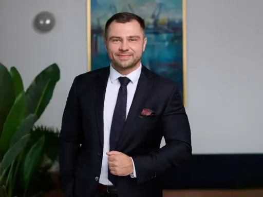 Mężczyzna w garniturze, stojący obok rośliny, z obrazem w tle.