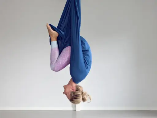 Kvinna hänger upp och ner i en blå tyghängmatta, utövar flying yoga.