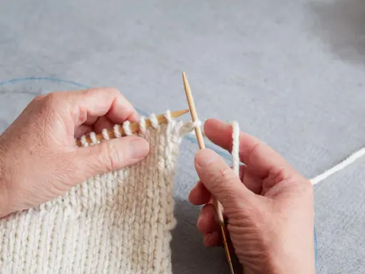 Händer stickar en vit resår med trästickor. En blå kabel syns i bakgrunden.