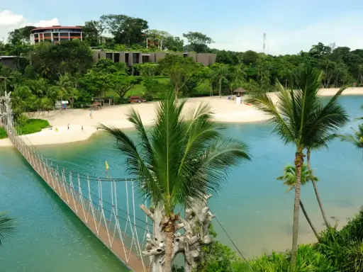 Hängende Brücke über türkisfarbenem Wasser führt zu einem sonnigen singapur strand mit Palmen und Resorts im Hintergrund.