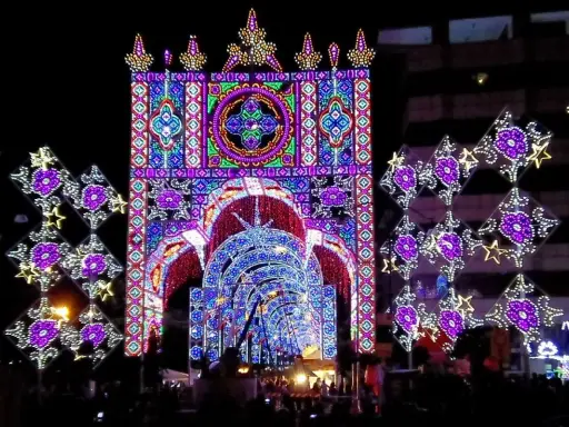 Un tunnel di luci colorate e decorazioni luminose, tipiche delle feste patronali, crea un'atmosfera magica e festosa.