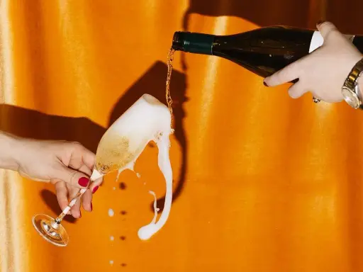 Schaumiges Sektglas läuft über, während eine Flasche auf orangefarbenem Hintergrund eingegossen wird. Eine Szene, die an die **zero dosage definition** erinnert, wo jeder Tropfen zählt.