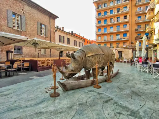 Statua di rinoceronte in una barca, in una piazza di Rimini, dove è nato Fellini. Tavolini all'aperto e palazzi colorati sullo sfondo.