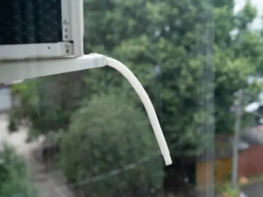 Tubo bianco flessibile per lo scarico condensa condizionatore, sporge da un'unità esterna, con alberi verdi sullo sfondo.