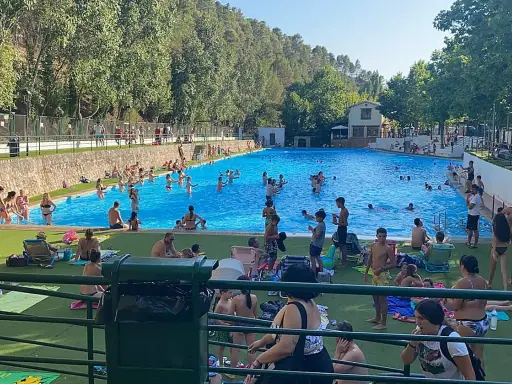 Gente disfrutando de un día soleado en la **piscina segura de la sierra**. Niños y adultos se refrescan en el agua azul, rodeados de árboles y un paisaje montañoso.