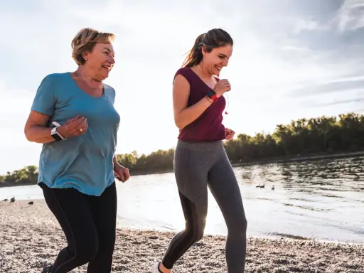 Zwei Frauen joggen am Wasser, um ihre Fettverbrennung anzuregen. Sie lächeln und genießen die Bewegung.