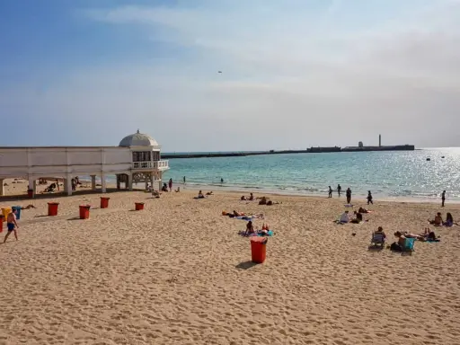 Sonniger Cadiz Strand mit Menschen, die sich am Meer entspannen. Ein weißes Gebäude mit Kuppel steht am Ufer.
